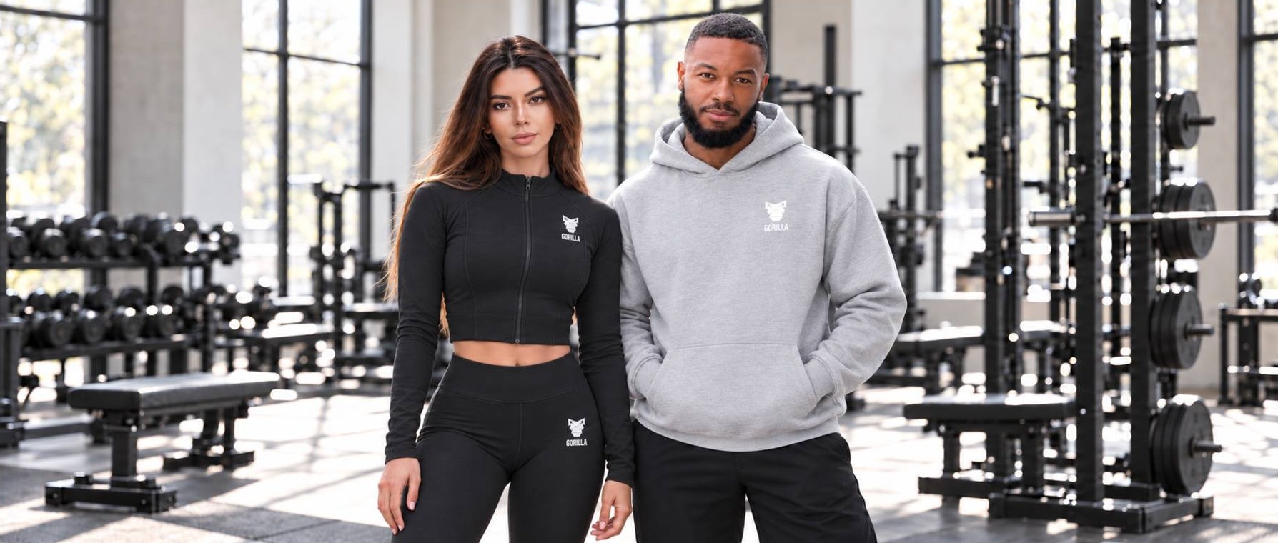 A woman in a black Gorilla zip jacket and leggings stands beside a man in a light grey Gorilla hoodie in a gym
